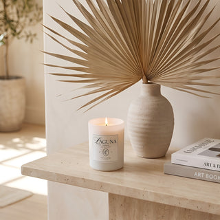 Panel 1 : Lit Laguna “Tide & Tuberose” candle on a textured beige travertine stone console table, next to a light-colored textured vase holding a dried palm leaf frond, and a stack of two books, with a potted olive tree in the background.