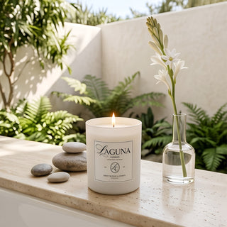 Panel 1 : Lit Laguna “Driftwood & Dawn” candle on a beige stone ledge next to a glass vase with a white tuberose flower, smooth grey stones, and a background of lush green potted ferns.