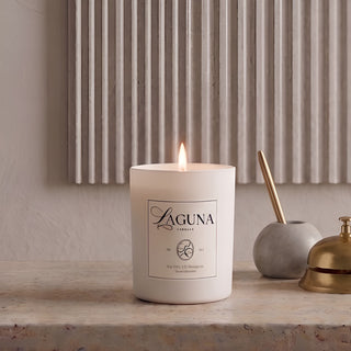 Panel 1: Lit Laguna “Sandalwood Shores” candle on a textured beige travertine stone console table, next to a grey marble pen holder with a gold pen and a brass desk bell, against a vertically fluted wall panel.