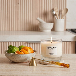 Panel 1: Lit Laguna candle displayed on a light marble kitchen countertop, accompanied by a ceramic bowl of fresh citrus fruits and candle accessories.
