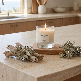Panel 1: Lit Laguna candle displayed on a light stone kitchen countertop, placed on a wooden board and styled with natural driftwood and dried lichen.