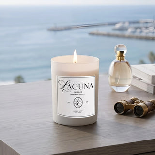 Panel 1: Lit Laguna “Harbor Mist” candle on a wooden surface with a glass perfume bottle and vintage binoculars overlooking a harbor view.
