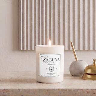 Panel 1: Lit Laguna “Sandalwood Shores” candle on a textured beige travertine stone console table, next to a grey marble pen holder with a gold pen and a brass desk bell, against a vertically fluted wall panel.