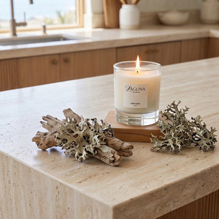 Panel 1: Lit Laguna candle displayed on a light stone kitchen countertop, placed on a wooden board and styled with natural driftwood and dried lichen.
