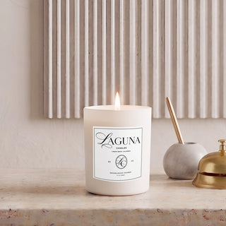Panel 1: Lit Laguna “Sandalwood Shores” candle on a textured stone console table against a fluted wall panel, next to a small grey ceramic holder and a brass bell.   