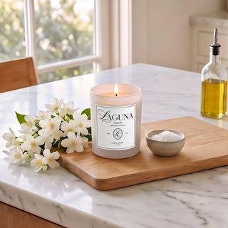 Panel 1: Lit Laguna “Laguna Breeze” candle on a wooden cutting board on a white marble kitchen countertop next to white flowers, a bowl of sea salt, and a glass oil dispenser[cite: 1].