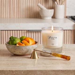 Panel 1: Lit Laguna candle displayed on a light marble kitchen countertop, accompanied by a ceramic bowl of fresh citrus fruits and candle accessories.