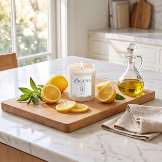 Panel 1: Lit Laguna “Citrus Cove” candle on a wooden cutting board on a white marble kitchen countertop next to fresh halved lemons, green sprigs, a glass oil dispenser, and a folded napkin.   