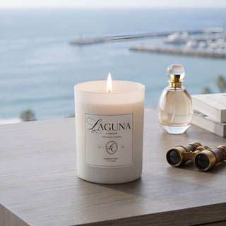Panel 1: Lit Laguna “Harbor Mist” candle on a wooden surface with a glass perfume bottle and vintage binoculars overlooking a harbor view.