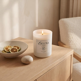 Panel 1 : Lit Laguna “Tide & Tuberose” candle on a light oak wood desk or table, next to a bowl of dried citrus slices and a smooth river stone, with a textured cream armchair in the background.