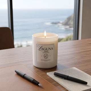 Panel 1: Lit Laguna “Driftwood & Dawn” candle on a leather desk blotter next to a beige notebook and two black pens, with a large window overlooking a blue ocean and coastal cliffs.