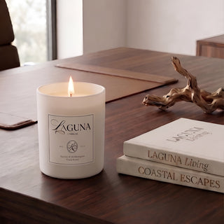 Panel 1: Lit Laguna “Sandalwood Shores” candle on a dark, rich walnut wood executive desk, next to a natural driftwood sculpture and a stack of two books (“LAGUNA Living” and “COASTAL ESCAPES”), with a hint of an office chair and window.