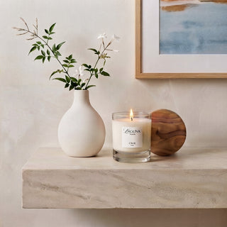 Panel 1: Lit Laguna candle placed on a minimalist cream-colored console table, styled with a white ceramic vase, delicate green branches, and coastal-inspired wall art.