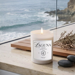 Panel 1: Lit Laguna “Harbor Mist” candle on a stone ledge with dark smooth stones and a wooden tray with foliage against an ocean background.