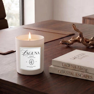 Panel 1: Lit Laguna “Sandalwood Shores” candle on a dark, rich walnut wood executive desk, next to a natural driftwood sculpture and a stack of two books (“LAGUNA Living” and “COASTAL ESCAPES”), with a hint of an office chair and window.