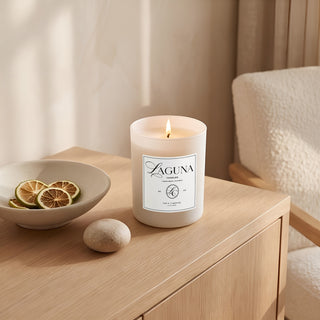 Panel 1: Lit Laguna “Tide & Tuberose” candle on a travertine console table next to a textured ceramic vase with a large dried palm leaf fan and stacked art books.   