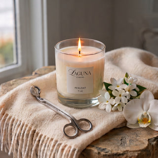 Panel 1: Lit Laguna “PERLOAT” candle resting on a soft, cream-colored knitted throw blanket, white flowers and a silver wick trimmer.