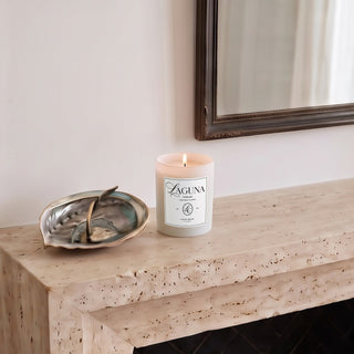 Panel 1: Lit Laguna “Laguna Breeze” candle on a textured stone fireplace mantel next to a decorative metallic seashell sculpture[cite: 1].