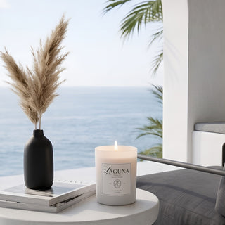 Panel 1: Lit Laguna “Harbor Mist” candle on a white round side table next to a stack of books, a black vase with pampas grass, and an ocean view with palm fronds.