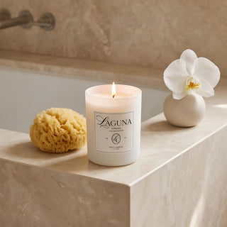 Panel 1 : Lit Laguna “Tide & Tuberose” candle on a polished beige marble bathroom vanity ledge, next to a natural sea sponge and a white vase with an orchid bloom, against a marble tile wall.
