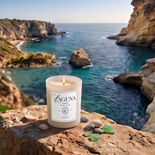 Panel 1: Lit Laguna “Laguna Breeze” candle on a rocky coastal cliffside ledge with smooth pebbles, sea glass, overlooking the vast blue ocean with sea stacks[cite: 1].