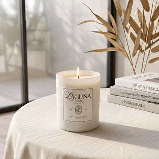 Panel 1: Lit Laguna “Driftwood & Dawn” candle on a textured beige linen tablecloth next to a stack of three “DESIGN” books and a window overlooking an ocean view with palm fronds.