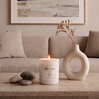 Panel 1: Lit Laguna “Sandalwood Shores” candle on a polished travertine coffee table, next to a cluster of smooth grey river stones and a distinctive white donut-shaped ceramic vase holding a single floral stem, in front of a linen sofa and abstract wall art.