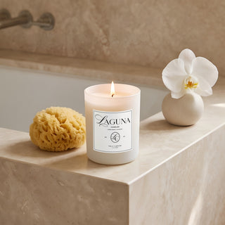 Panel 1: Lit Laguna “Tide & Tuberose” candle on a stone bathtub ledge next to a natural sea sponge and a small vase with a white orchid blossom. 