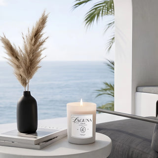 Panel 1: Lit Laguna “Harbor Mist” candle on a white round side table next to a stack of books, a black vase with pampas grass, and an ocean view with palm fronds.