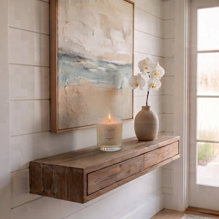 Panel 1: Lit Laguna “PERLOAT” candle displayed on a floating wooden wall shelf beneath a textured coastal art piece and a vase of white orchids.