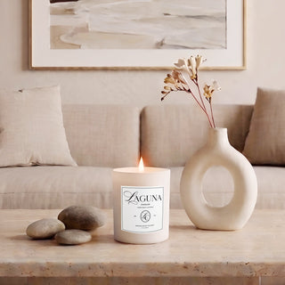 Panel 1: Lit Laguna “Sandalwood Shores” candle on a polished travertine coffee table, next to a cluster of smooth grey river stones and a distinctive white donut-shaped ceramic vase holding a single floral stem, in front of a linen sofa and abstract wall art.