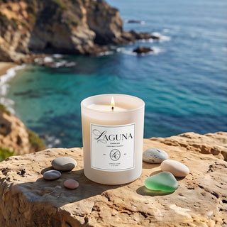 Panel 1: Lit Laguna “Citrus Cove” candle on a rocky coastal cliffside ledge with smooth pebble stones, sea glass, overlooking a vibrant blue ocean view.   