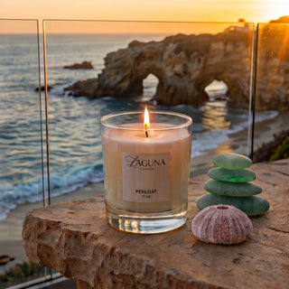 Panel 1: Lit Laguna PERLOAT candle on an outdoor stone ledge with stacked sea glass and a sea urchin shell against the ocean sunset.