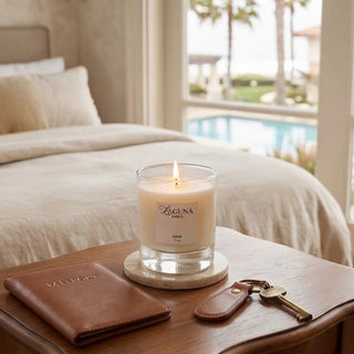 Panel 1: Lit Laguna candle placed on a wooden bedside table in a bright bedroom, accompanied by a leather passport holder, key case, and keys, with a peaceful poolside view in the background.