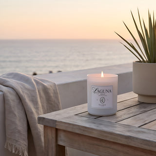 Panel 1 : Lit Laguna “Tide & Tuberose” candle on a weathered wooden slat side table on an outdoor balcony, next to a succulent plant in a grey planter and a draped grey throw blanket, overlooking the ocean at sunset.