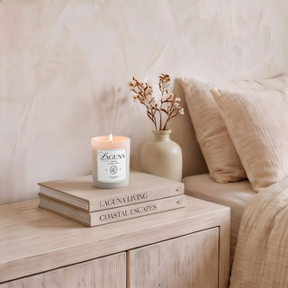 Panel 1: Lit Laguna “Laguna Breeze” candle on a light wooden bedside table next to stacked books (“Laguna Living” and “Coastal Escapes”) and a vase with dried foliage[cite: 1].