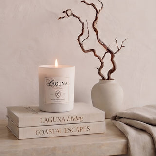 Panel 1: Lit Laguna “Sandalwood Shores” candle on a cream-colored marble ledge, next to a textured cream vase with a twisted branch sculpture and a draped beige linen throw, against a textured beige wall.