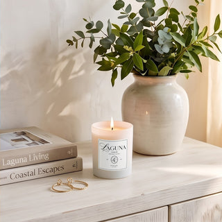 Panel 1: Lit Laguna “Citrus Cove” candle on a light wooden surface next to stacked books, gold hoop rings, and a ceramic vase filled with fresh eucalyptus branches.   
