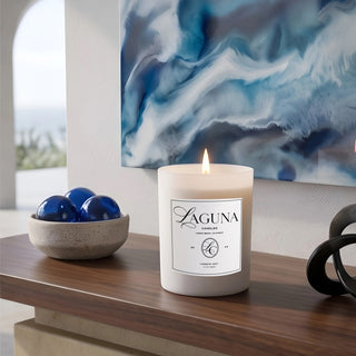 Panel 4: Lit Laguna “Harbor Mist” candle on a wooden console table next to a bowl of blue glass spheres and an abstract blue wall art piece.