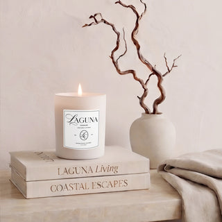 Panel 1: Lit Laguna “Sandalwood Shores” candle on a cream-colored marble ledge, next to a textured cream vase with a twisted branch sculpture and a draped beige linen throw, against a textured beige wall.