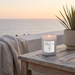 Panel 1: Lit Laguna “Tide & Tuberose” candle on a wooden outdoor balcony table with a potted plant, overlooking an ocean sunset view with a draped throw blanket. 