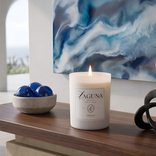 Panel 4: Lit Laguna “Harbor Mist” candle on a wooden console table next to a bowl of blue glass spheres and an abstract blue wall art piece.