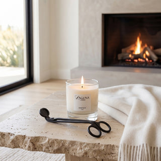 Panel 1: Lit Laguna candle placed on a natural stone table beside a soft cream blanket, with a lit fireplace in the background creating a warm indoor setting.