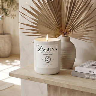 Panel 1 : Lit Laguna “Tide & Tuberose” candle on a textured beige travertine stone console table, next to a light-colored textured vase holding a dried palm leaf frond, and a stack of two books, with a potted olive tree in the background.