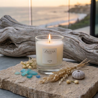 Panel 1: Lit Laguna “Layne” candle on a stone surface with driftwood, sea glass, and dried wheat.
