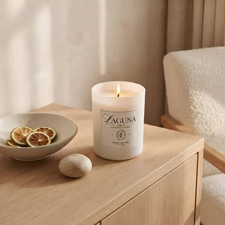Panel 1 : Lit Laguna “Tide & Tuberose” candle on a light oak wood desk or table, next to a bowl of dried citrus slices and a smooth river stone, with a textured cream armchair in the background.