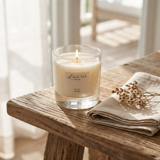 Panel 1: Lit Laguna “821-A” candle on a wooden table with a folded cream cloth and a small dried floral stem.
