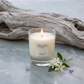 Panel 1: Lit Laguna “Forest Ave.” candle with driftwood and small white and purple flowers on a marble surface.